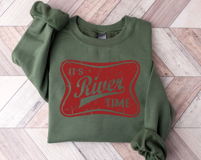 May include: An olive green crewneck sweatshirt featuring a distressed red rectangular graphic with rounded corners. The text "IT'S River TIME" is printed in olive green across the center of the red graphic, matching the color of the sweatshirt fabric.