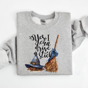 May include: A heather gray crewneck sweatshirt featuring a graphic print of a blue witch hat with autumn leaves and a wooden broomstick with a blue ribbon. The text reads, "Yes, I can drive a stick!" in a black, stylized cursive font.