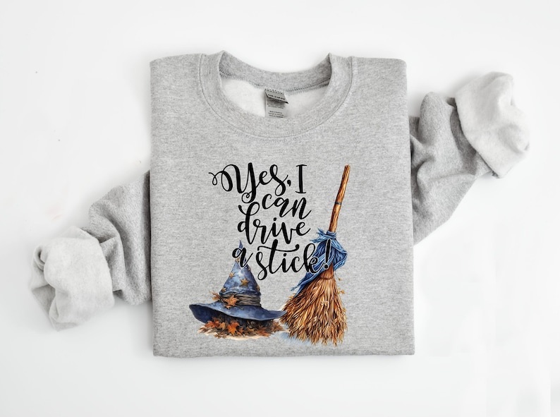 May include: A heather gray crewneck sweatshirt featuring a graphic print of a blue witch hat with autumn leaves and a wooden broomstick with a blue ribbon. The text reads, "Yes, I can drive a stick!" in a black, stylized cursive font.