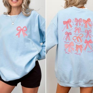 May include: A light blue crewneck sweatshirt featuring a small pink bow graphic on the front left chest. The back displays a large collection of pink bows in various styles, accompanied by cursive text including words like loved, precious, and strong.