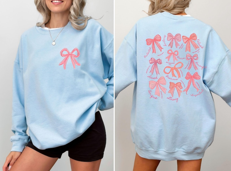May include: A light blue crewneck sweatshirt featuring a small pink bow graphic on the front left chest. The back displays a large collection of pink bows in various styles, accompanied by cursive text including words like loved, precious, and strong.