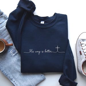 May include: A navy blue sweatshirt with the text "His way is better" and a cross design. The sweatshirt is paired with a pair of light blue jeans, a brown mug with coffee, and gray sneakers.