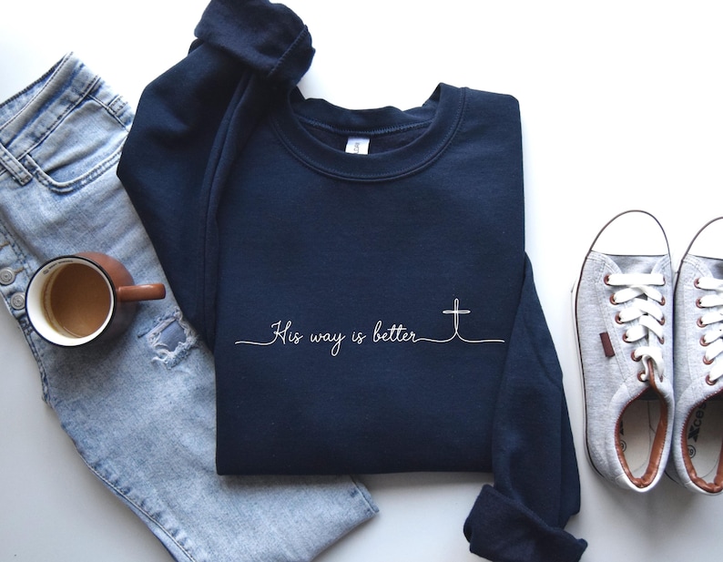May include: A navy blue sweatshirt with the text "His way is better" and a cross design. The sweatshirt is paired with a pair of light blue jeans, a brown mug with coffee, and gray sneakers.