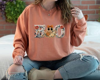 May include: A terracotta-colored crewneck sweatshirt featuring an embroidered graphic on the chest. The design spells out BOO, with the letter O replaced by a ghost figure wearing a bow, all decorated with colorful floral and leaf patterns.