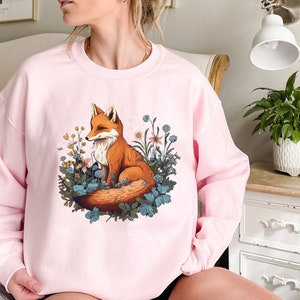 May include: A light pink crewneck sweatshirt featuring a central graphic illustration of an orange fox sitting amidst a cluster of blue and yellow wildflowers and green foliage. The design is printed on the front of the long-sleeved pullover garment.