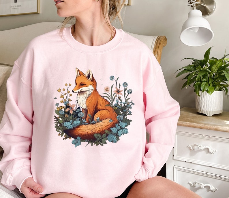 May include: A light pink crewneck sweatshirt featuring a central graphic illustration of an orange fox sitting amidst a cluster of blue and yellow wildflowers and green foliage. The design is printed on the front of the long-sleeved pullover garment.