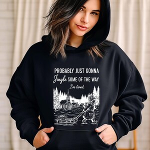 Puede incluir: Sudadera con capucha negra con el texto "Probably just gonna jingle some of the way, I'm tired." Un gr&aacute;fico en blanco y negro representa a una persona tirando de un trineo con regalos hacia una iglesia en una escena invernal.