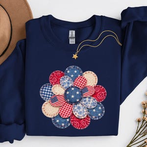May include: A navy blue sweatshirt with a floral design made from fabric patches in red, white, and blue patterns. The patches feature stars, stripes, and floral prints. A gold necklace with a star pendant is on the sweatshirt.