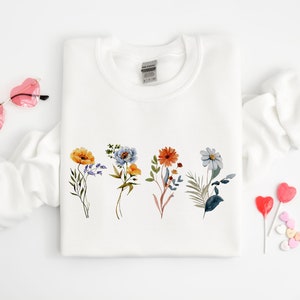 May include: White sweatshirt with a floral design featuring five different flowers in shades of blue, orange, yellow, and green. The flowers are arranged in a row across the centre of the sweatshirt.