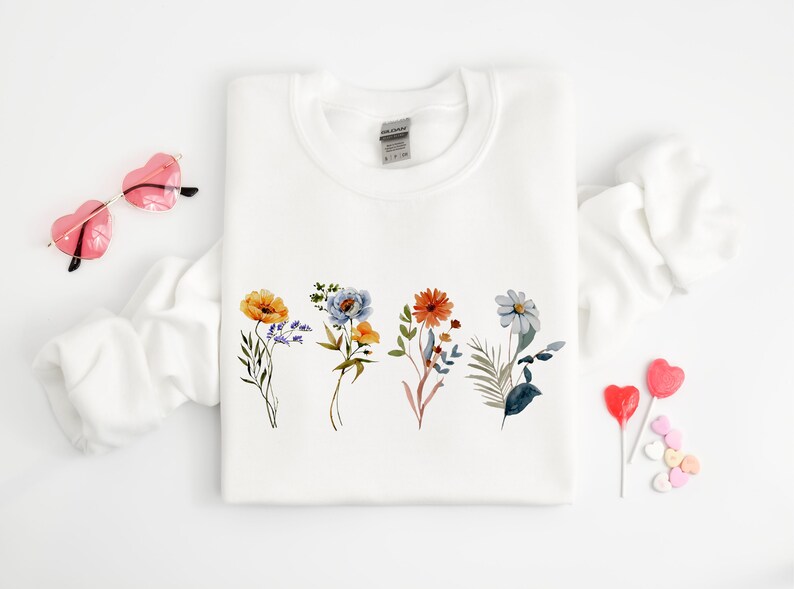May include: White sweatshirt with a floral design featuring five different flowers in shades of blue, orange, yellow, and green. The flowers are arranged in a row across the centre of the sweatshirt.