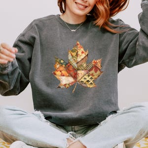 May include: A dark gray sweatshirt featuring a large, colorful maple leaf design. The leaf is composed of various fabric patterns in autumn colors like orange, yellow, and brown. The sweatshirt has long sleeves and a relaxed fit.