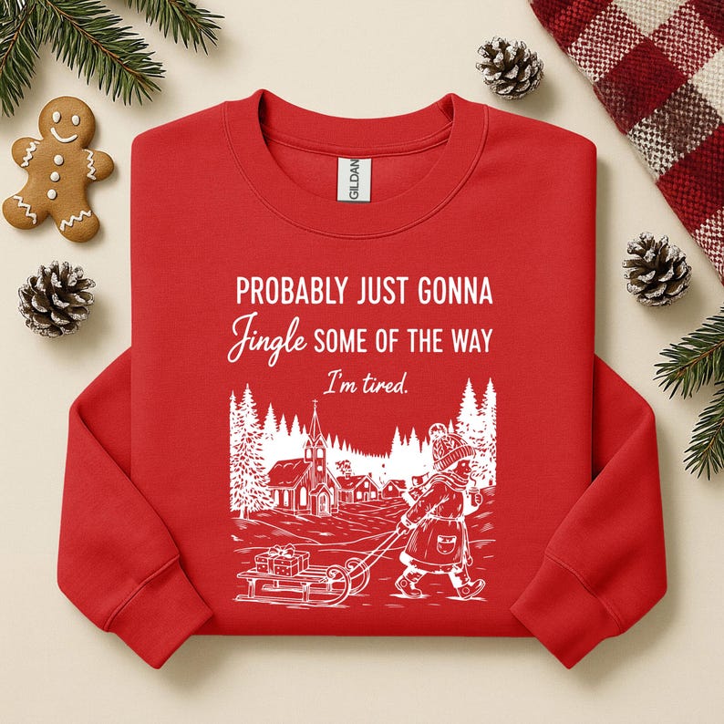 Puede incluir: Una sudadera roja con el texto "Probably Just Gonna Jingle Some of the Way, I'm tired." Un gr&aacute;fico blanco representa a un ni&ntilde;o tirando de un trineo por un pueblo nevado. La sudadera est&aacute; rodeada de decoraciones navide&ntilde;as.