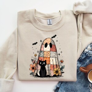 May include: A beige sweatshirt featuring a colorful patchwork ghost design with a black cat, flowers, and bats. The graphic is in the center of the sweatshirt. The sweatshirt is paired with blue jeans and a coffee mug.