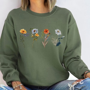 May include: A green sweatshirt with a floral print featuring orange, yellow, blue, and white flowers. The flowers are arranged in a row across the chest of the sweatshirt.