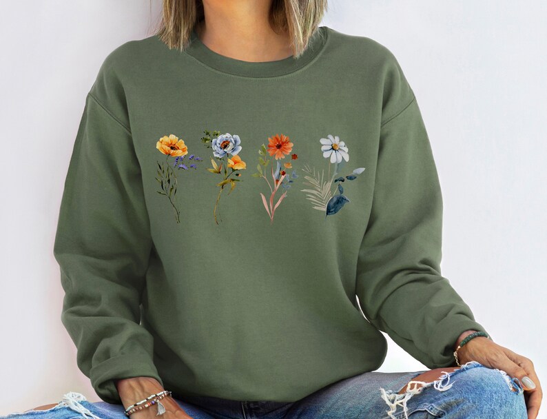May include: A green sweatshirt with a floral print featuring orange, yellow, blue, and white flowers. The flowers are arranged in a row across the chest of the sweatshirt.