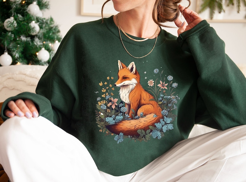 May include: A dark green crewneck sweatshirt featuring a central graphic illustration of an orange and white fox sitting amidst a cluster of blue, pink, and yellow wildflowers and green foliage. The sweatshirt has long sleeves and a ribbed neckline.