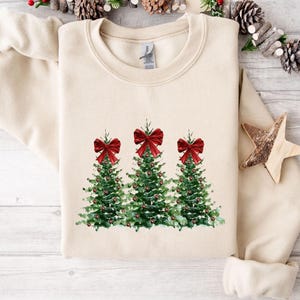 Pode incluir: Um moletom creme com um design em aquarela de três árvores de Natal. Cada árvore tem um laço vermelho no topo e é decorada com bagas vermelhas. O moletom está sobre uma superfície de madeira clara.