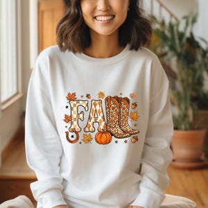 May include: A white long-sleeve sweatshirt featuring a graphic print that spells the word FALL. The letters are decorated with a checkered pattern, cactus motifs, and leopard print. The design includes a pumpkin, a horseshoe, and autumn leaves.