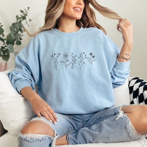 May include: A light blue crewneck sweatshirt featuring a black line art graphic of five distinct wildflowers across the chest. The long-sleeve pullover has a ribbed collar, cuffs, and hem, providing a relaxed fit with a simple botanical design.
