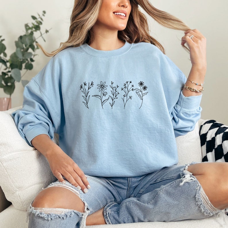 May include: A light blue crewneck sweatshirt featuring a black line art graphic of five distinct wildflowers across the chest. The long-sleeve pullover has a ribbed collar, cuffs, and hem, providing a relaxed fit with a simple botanical design.