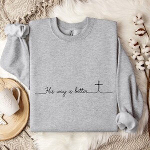 May include: A light gray sweatshirt with the text "His way is better" and a cross graphic in black. The long-sleeved crew neck shirt is displayed on a white fur-like surface.