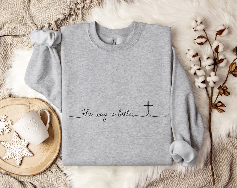 May include: A light gray sweatshirt with the text "His way is better" and a cross graphic in black. The long-sleeved crew neck shirt is displayed on a white fur-like surface.