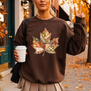 May include: A brown sweatshirt featuring a large, colorful leaf design made of patchwork fabrics. The leaf design incorporates various patterns and colors, including orange, red, green, and yellow. The sweatshirt is a solid brown color.