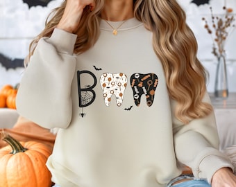 May include: Beige sweatshirt with the word "BOO" in black letters and two tooth designs. The "B" has a spider web and spider, and the teeth are decorated with Halloween candy. Small bats are scattered around the letters.