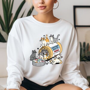 K&ouml;nnte beinhalten: Ein wei&szlig;es Langarm-Sweatshirt mit einer Grafik einer gelben Pillendose, beschriftet mit Serotonin Booster und dem Nebeneffekt Cute Aggression. Das Design zeigt mehrere bunte Cartoon-Katzen, die mit der Dose und den Pillen interagieren.