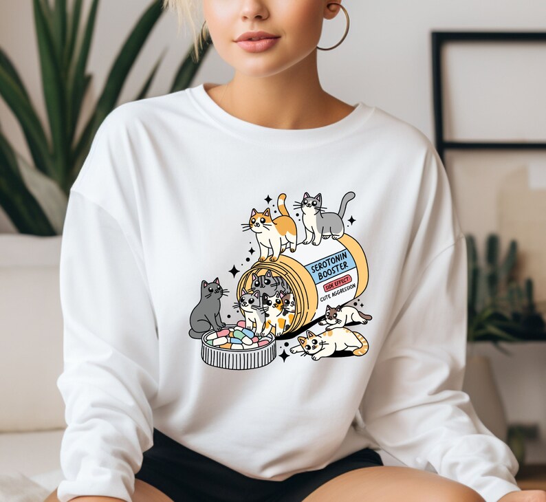 K&ouml;nnte beinhalten: Ein wei&szlig;es Langarm-Sweatshirt mit einer Grafik einer gelben Pillendose, beschriftet mit Serotonin Booster und dem Nebeneffekt Cute Aggression. Das Design zeigt mehrere bunte Cartoon-Katzen, die mit der Dose und den Pillen interagieren.