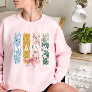 Pu&ograve; includere: Felpa rosa con un design floreale e la parola "MAMA" in lettere bianche.