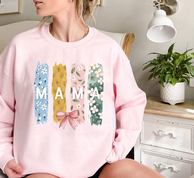 Pu&ograve; includere: Felpa rosa con un design floreale e la parola "MAMA" in lettere bianche.