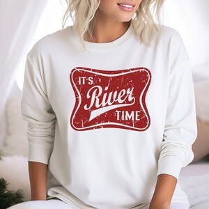 May include: A cream-colored long-sleeve sweatshirt featuring a distressed red rectangular graphic with rounded corners. The graphic contains the white text "IT'S River TIME" in a stylized font, with a weathered texture throughout the design.