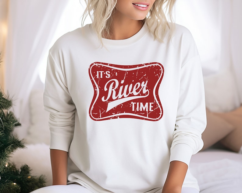May include: A cream-colored long-sleeve sweatshirt featuring a distressed red rectangular graphic with rounded corners. The graphic contains the white text "IT'S River TIME" in a stylized font, with a weathered texture throughout the design.