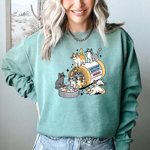 K&ouml;nnte beinhalten: Ein salbeigr&uuml;nes Sweatshirt mit Rundhalsausschnitt und einer Grafik einer Pillendose mit der Aufschrift Serotonin Booster sowie dem Text Side Effect Cute Aggression. Das Design zeigt mehrere Cartoon-Katzen, die mit der Dose und einem mit bunten Pillen gef&uuml;llten Deckel spielen.