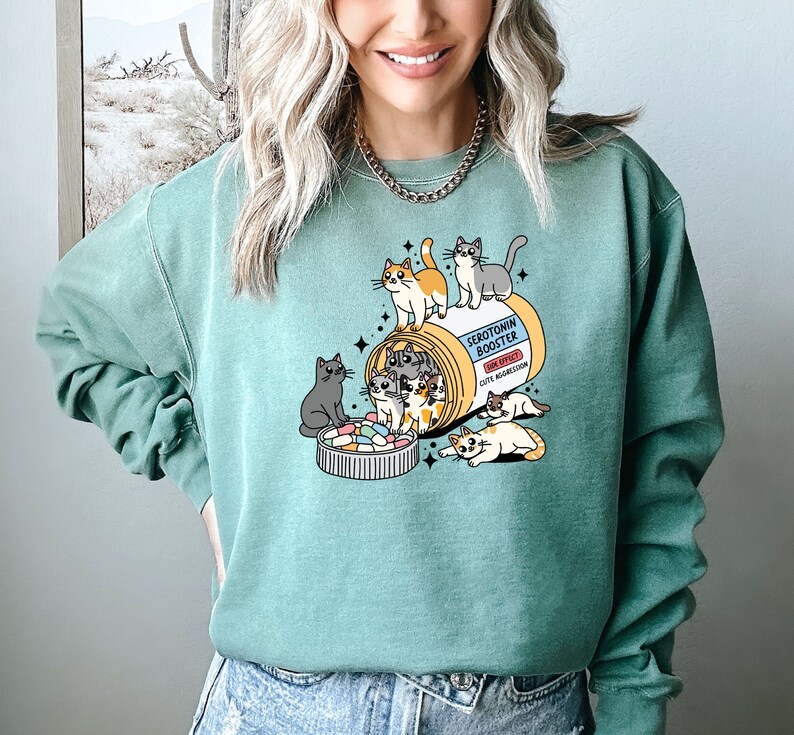 K&ouml;nnte beinhalten: Ein salbeigr&uuml;nes Sweatshirt mit Rundhalsausschnitt und einer Grafik einer Pillendose mit der Aufschrift Serotonin Booster sowie dem Text Side Effect Cute Aggression. Das Design zeigt mehrere Cartoon-Katzen, die mit der Dose und einem mit bunten Pillen gef&uuml;llten Deckel spielen.