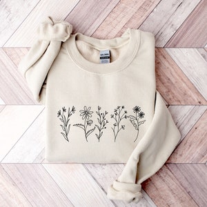 May include: A sand-colored crewneck sweatshirt featuring a black line art illustration of five distinct wildflowers across the chest. The garment has long sleeves with ribbed cuffs and a ribbed neckline, with a Gildan brand label visible inside.