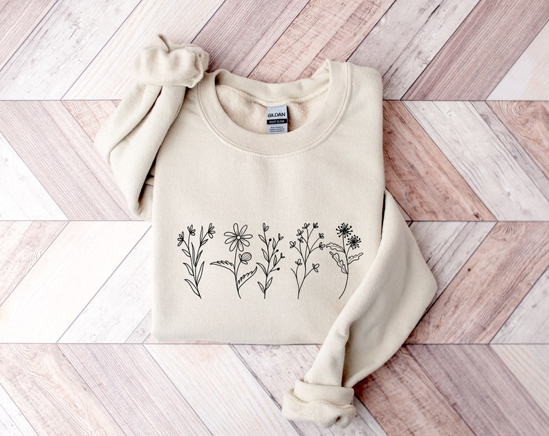 May include: A sand-colored crewneck sweatshirt featuring a black line art illustration of five distinct wildflowers across the chest. The garment has long sleeves with ribbed cuffs and a ribbed neckline, with a Gildan brand label visible inside.