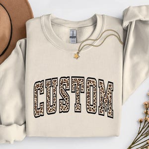 May include: Cream-colored sweatshirt with the word "CUSTOM" in a leopard print design. The text is outlined in black. A gold necklace with a star pendant is draped on the sweatshirt. A brown hat and flowers are in the background.