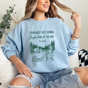 Puede incluir: Sudadera azul claro con el texto "Probably Just Gonna Jingle Some of the Way, I'm tired." Una ilustraci&oacute;n verde muestra a una persona tirando de un trineo por un pueblo nevado. La sudadera es de estilo casual.