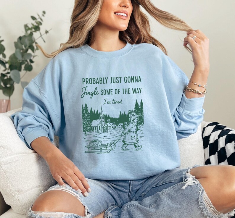 Puede incluir: Sudadera azul claro con el texto "Probably Just Gonna Jingle Some of the Way, I'm tired." Una ilustraci&oacute;n verde muestra a una persona tirando de un trineo por un pueblo nevado. La sudadera es de estilo casual.