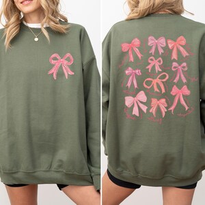 May include: An olive green crewneck sweatshirt featuring a single pink bow graphic on the upper left chest and a large collection of various pink bow illustrations printed on the back, each accompanied by small cursive text labels below the bows.