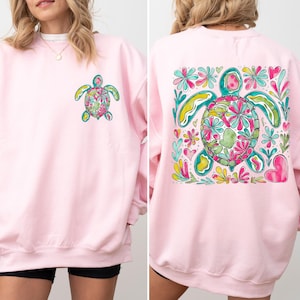 May include: A light pink crewneck sweatshirt featuring a small, colorful watercolor-style sea turtle graphic on the left chest and a large, matching sea turtle design surrounded by vibrant floral and heart motifs on the back of the garment.