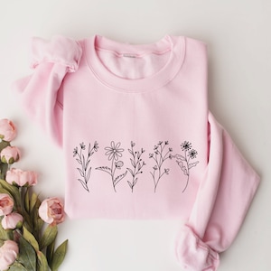 May include: A light pink crewneck sweatshirt featuring a black line art illustration of five distinct wildflowers across the center. The garment has long sleeves with ribbed cuffs and a ribbed neckline, providing a simple and clean aesthetic.