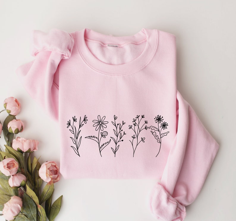May include: A light pink crewneck sweatshirt featuring a black line art illustration of five distinct wildflowers across the center. The garment has long sleeves with ribbed cuffs and a ribbed neckline, providing a simple and clean aesthetic.