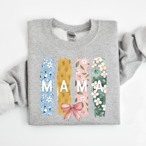 Pu&ograve; includere: Una felpa grigia con un motivo floreale e la parola "MAMA" in lettere bianche. Il motivo floreale &egrave; in tonalit&agrave; di blu, giallo, rosa e verde.