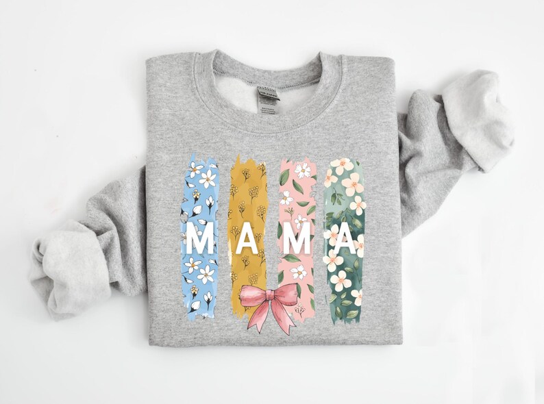 Pu&ograve; includere: Una felpa grigia con un motivo floreale e la parola "MAMA" in lettere bianche. Il motivo floreale &egrave; in tonalit&agrave; di blu, giallo, rosa e verde.