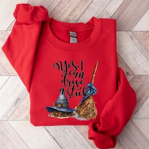 May include: A bright red long-sleeve sweatshirt featuring a graphic print of a blue witch hat and a straw broom. The text "Yes, I can drive a stick!" is printed in black cursive lettering above the hat and broom illustrations on the front.