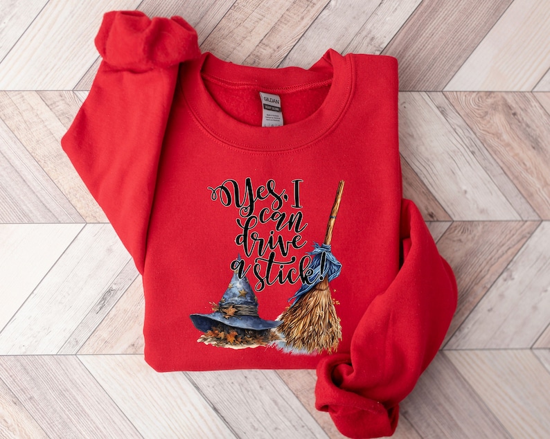 May include: A bright red long-sleeve sweatshirt featuring a graphic print of a blue witch hat and a straw broom. The text "Yes, I can drive a stick!" is printed in black cursive lettering above the hat and broom illustrations on the front.