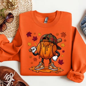 Op de afbeelding: Een oranje sweatshirt met een afbeelding van een pompoen in de vorm van een honkbal met stiksels, dragend een geruite honkbalpet en hoge sneakers. Het personage houdt een houten honkbalknuppel vast, omringd door vallende herfstbladeren.